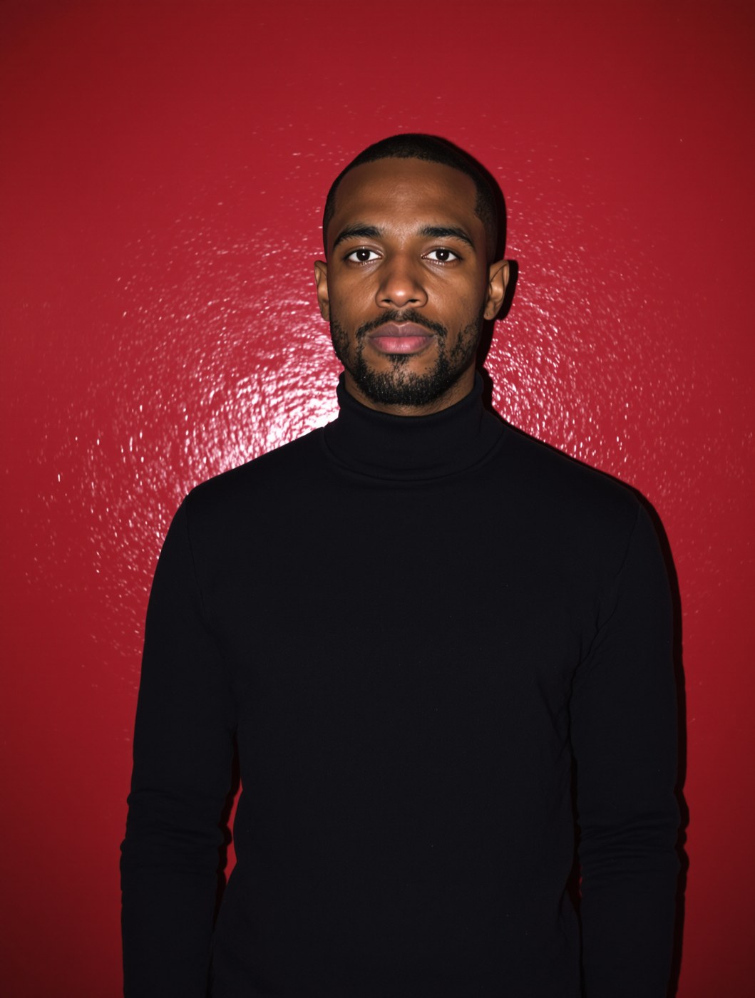 A man in a black turtleneck photographed with hard flash against a deep red wall.